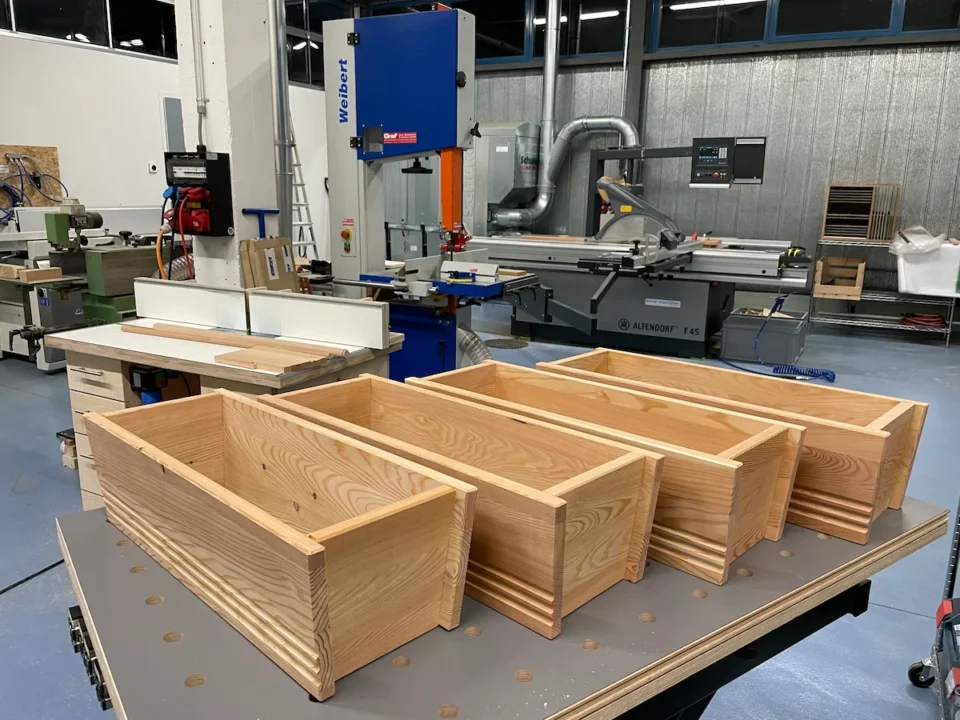 Blumenkisten aus Holz auf einem Tisch in einer Werkstatt mit verschiedenen Maschinen und Werkzeugen im Hintergrund.