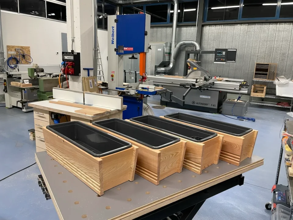 Blumenkisten aus Holz auf einem Arbeitstisch in einer modernen Werkstatt mit verschiedenen Maschinen und Werkzeugen im Hintergrund.
