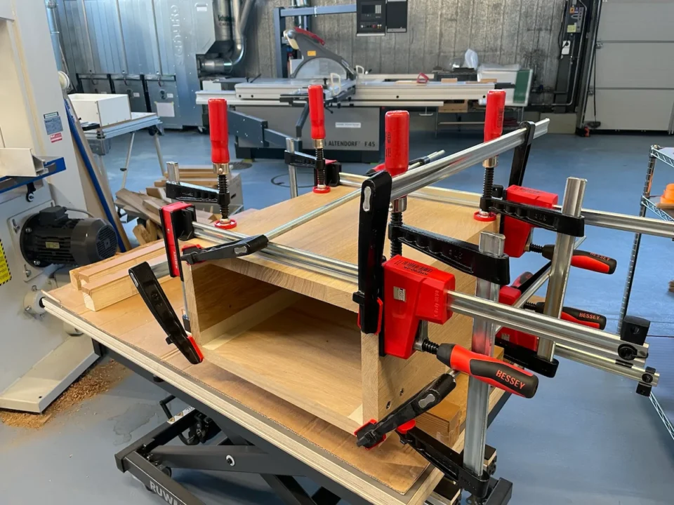 Zusammengespannte Holzbretter auf einer Werkbank in einer Werkstatt, im Hintergrund verschiedene Werkzeuge und Maschinen.