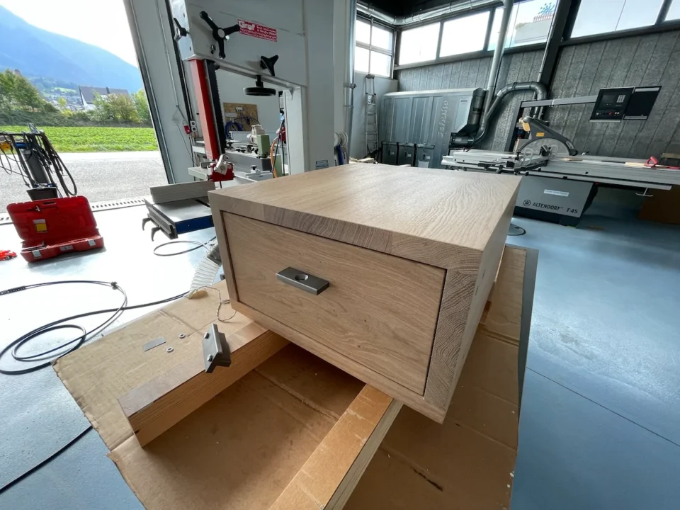 Eine grosse Holzschublade mit Stahlgriff auf einer Werkbank in einer gut ausgestatteten Werkstatt, im Hintergrund verschiedene Werkzeuge und Maschinen.