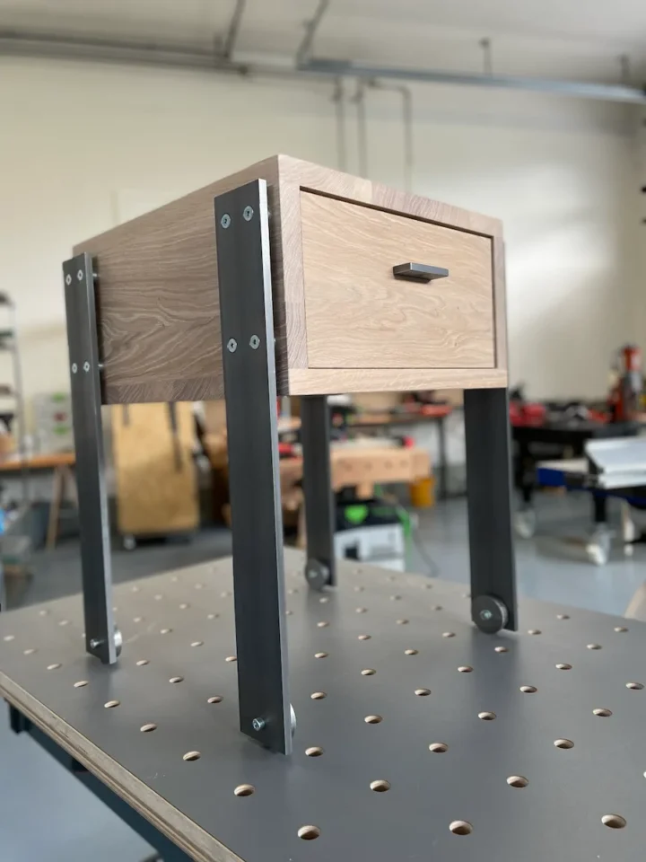 Ein handgefertigter Nachttisch aus Holz mit einer Schublade und Stahlbeinen, ausgestellt auf einer Werkbank in einer Werkstatt.