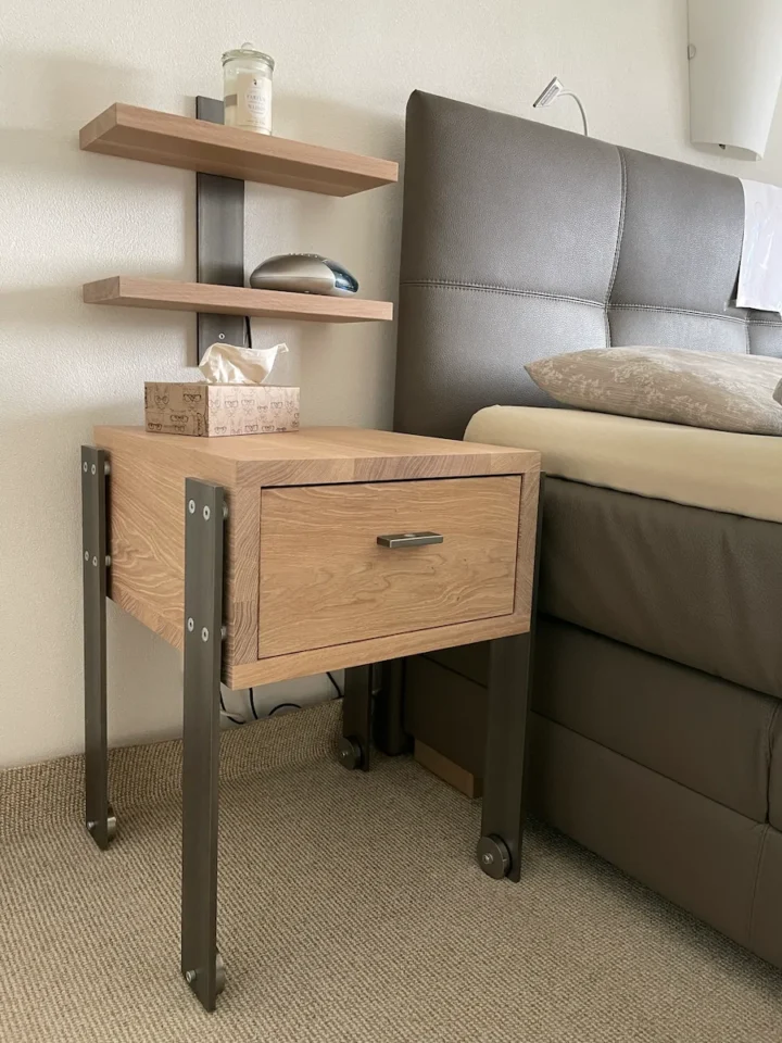 Ein hölzerner Nachttisch mit einer Schublade und Stahlbeinen, neben einem Bett mit grauer Polsterung, unter einem kleinen, an der Wand montierten Regal.