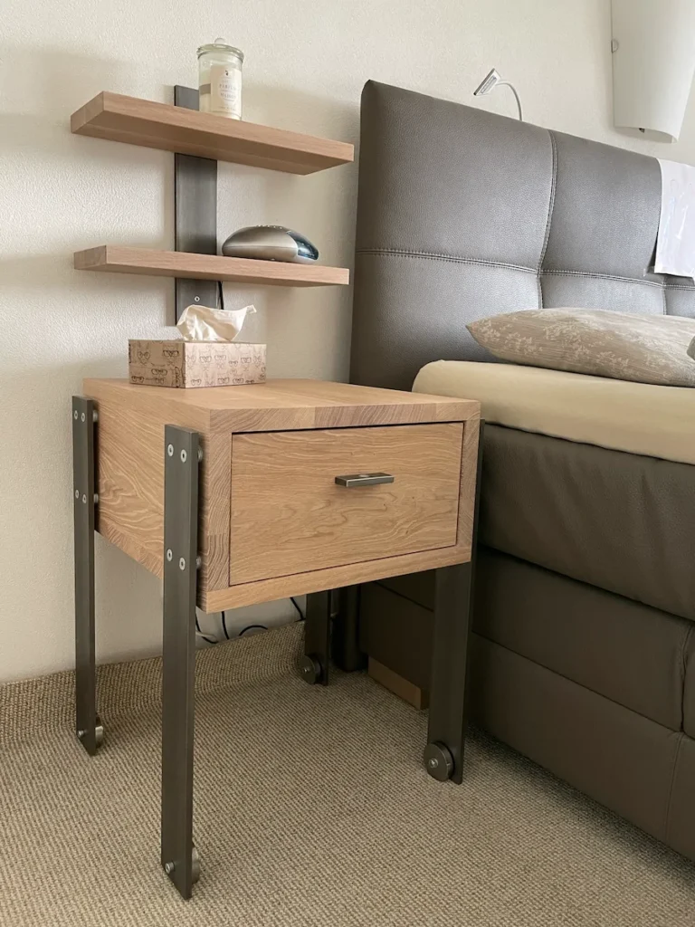 Ein hölzerner Nachttisch mit einer Schublade und Stahlbeinen, neben einem Bett mit grauer Polsterung, unter einem kleinen, an der Wand montierten Regal.