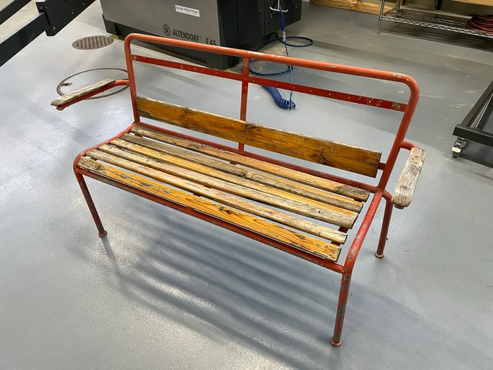 Eine verwitterte Gartenbank aus Holz und Metall mit einem abblätternden roten Rahmen in einer Werkstatt.