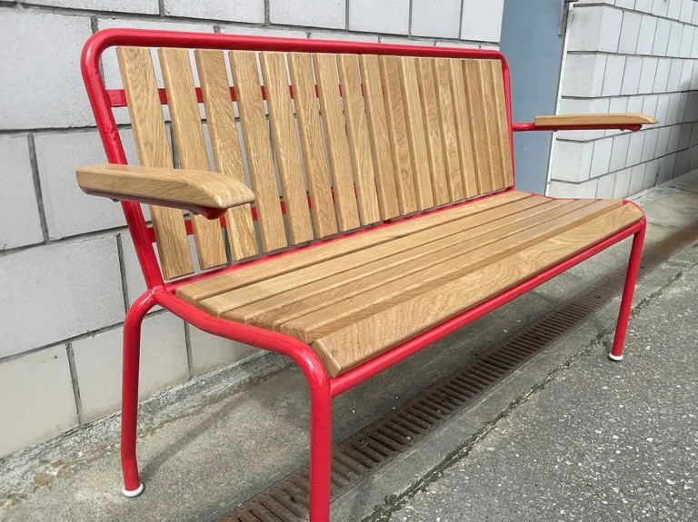Eine restaurierte Gartenbank aus Holz und rotem Metall mit Armlehnen vor einer grauen Betonwand.