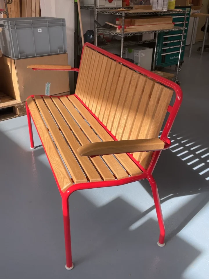 Eine restaurierte Gartenbank aus Holz und rotem Metall mit Armlehnen vom Sonnenlicht beleuchtet.