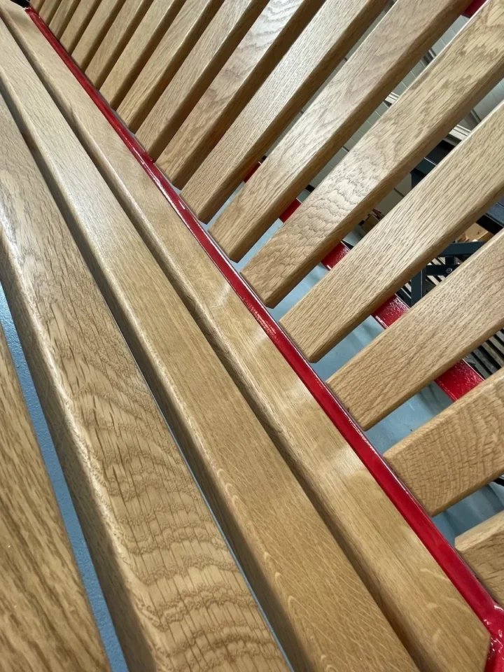 Nahaufnahme einer restaurierten Gartenbank aus Holzlatten mit einer horizontal zwischen den Latten verlaufenden roten Metallstange.