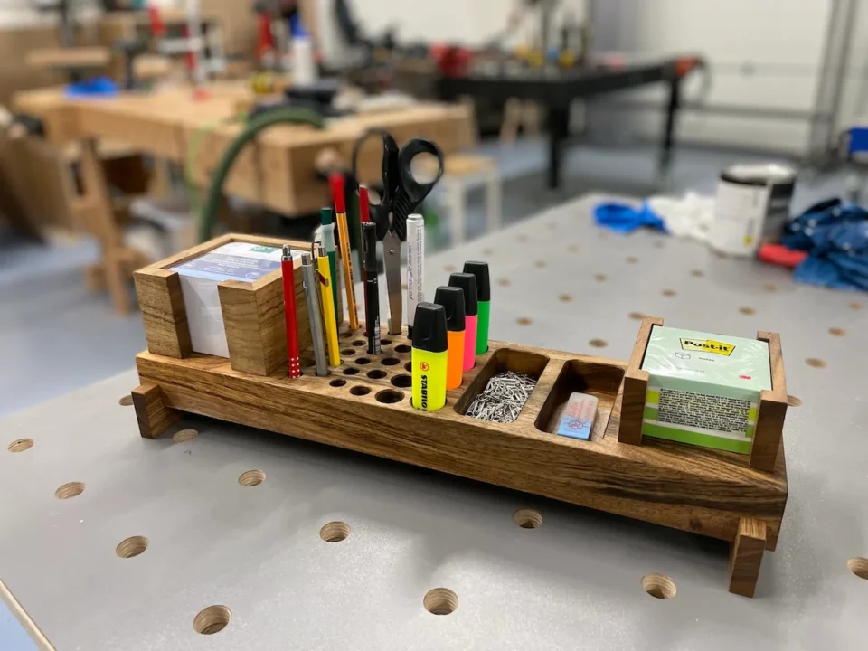 Ein Tischorganizer aus Holz mit verschiedenen Schreibwaren wie Stiften, Scheren und Notizblöcken, platziert auf einem Metallarbeitstisch in einer Werkstatt.